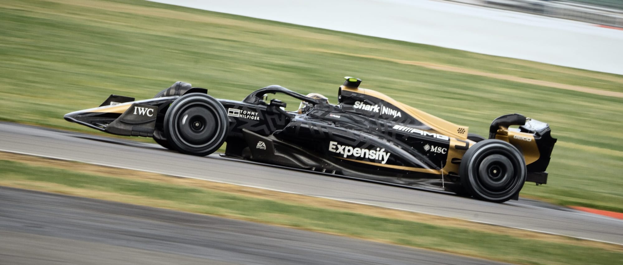 A black and gold Formula 1 style APXGP racing car driving at high speed on a track with motion blur background.