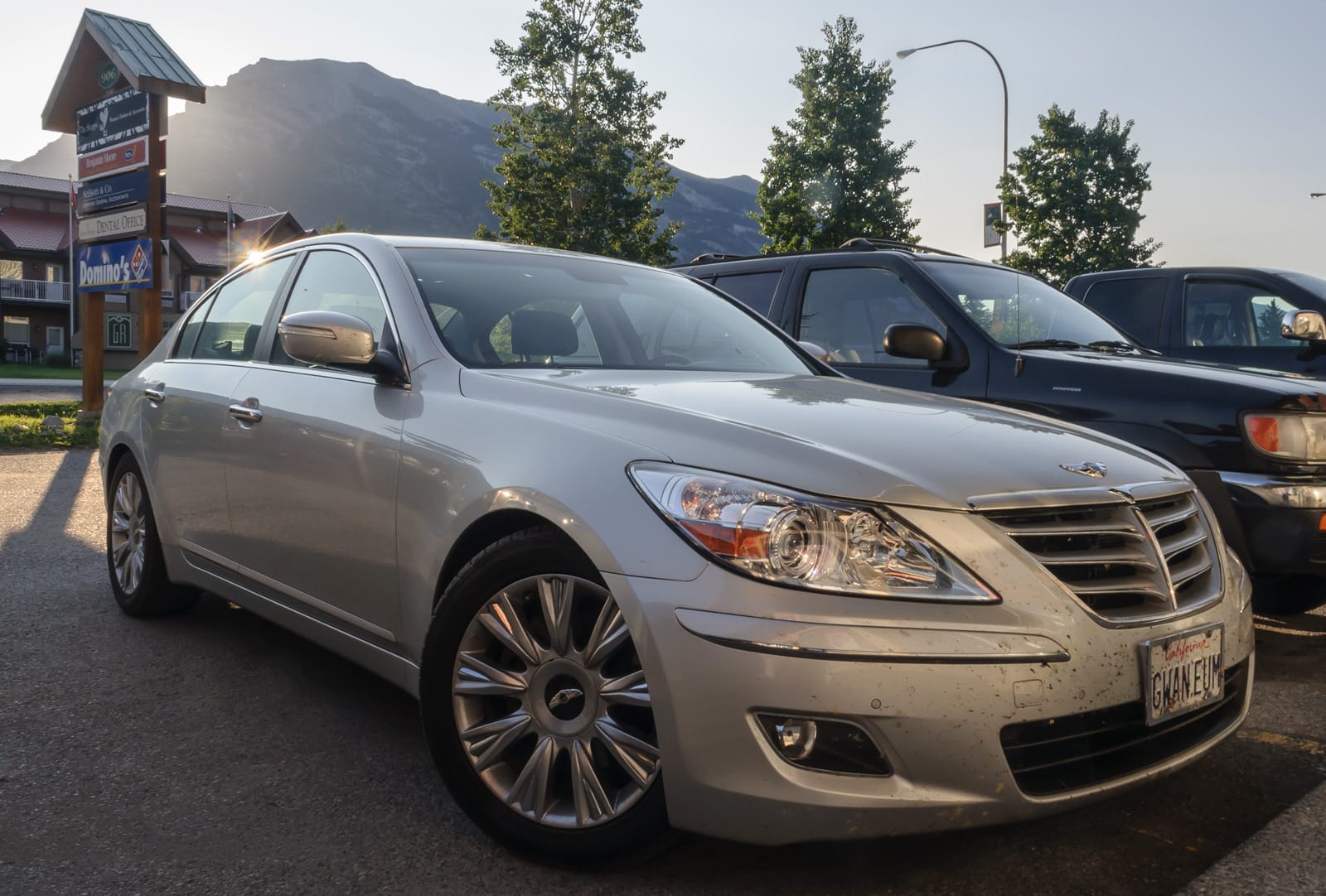 A silver Genesis sedan parked in an outdoor parking lot with mountain scenery and commercial signs visible in the background.