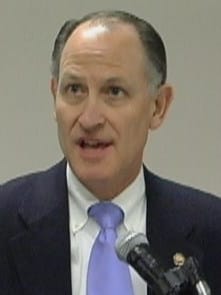 A close-up portrait of Jeffrey Kessler speaking into a microphone while wearing a suit and light blue tie.