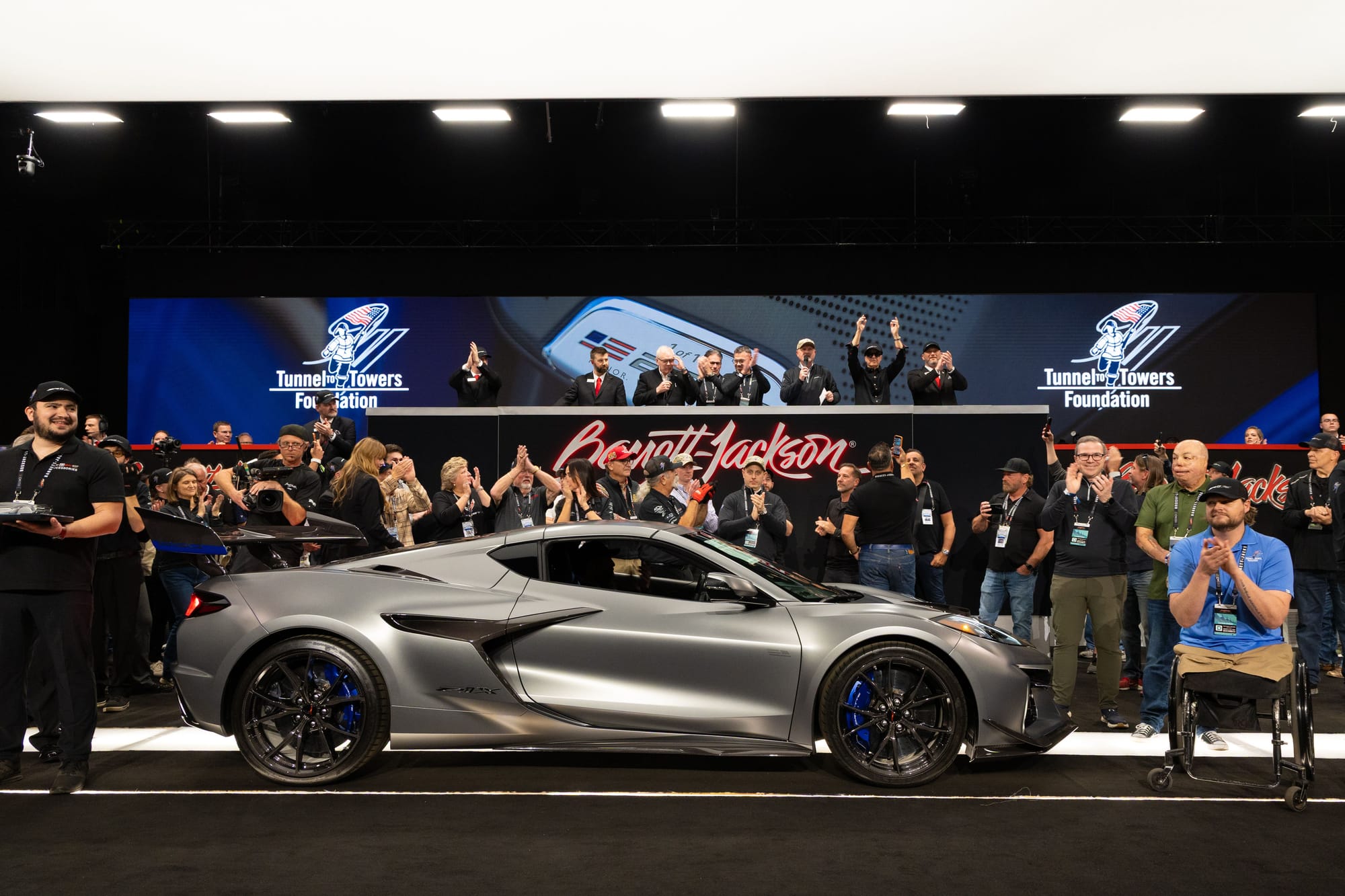 A matte gray Chevrolet Corvette C8 on stage at a Barrett-Jackson auction block surrounded by crowd members, auctioneers, and prominent Barrett-Jackson branding.