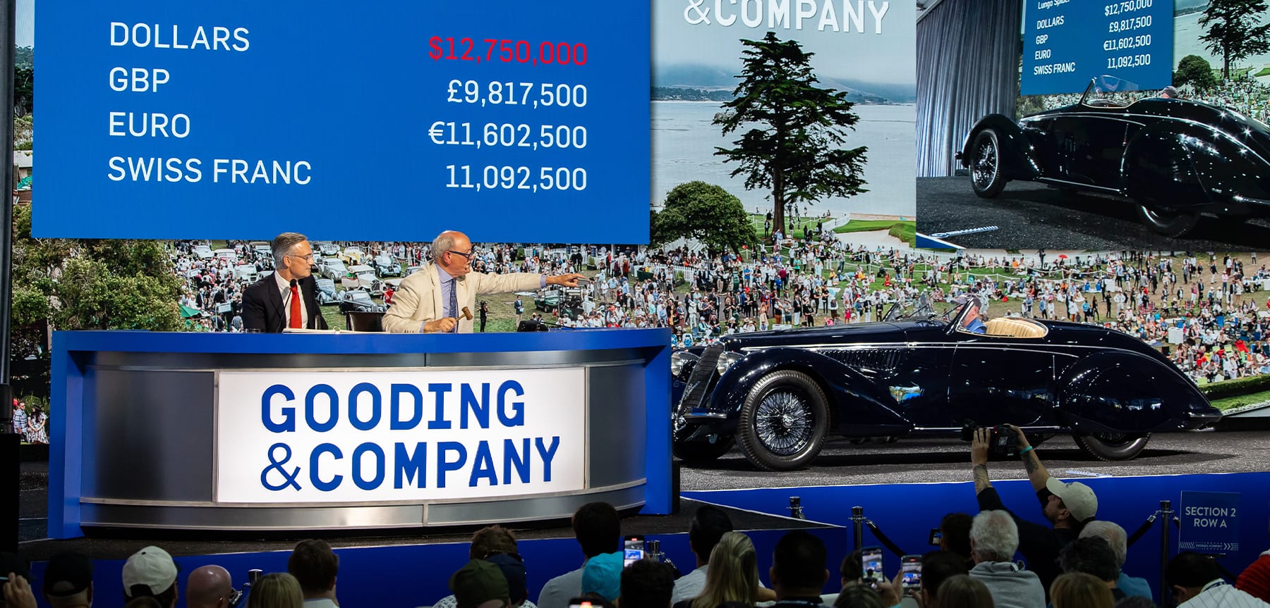 A Gooding & Company car auction event stage featuring an auctioneer at a branded podium, a high-value bid digital board, and a vintage classic car on display.