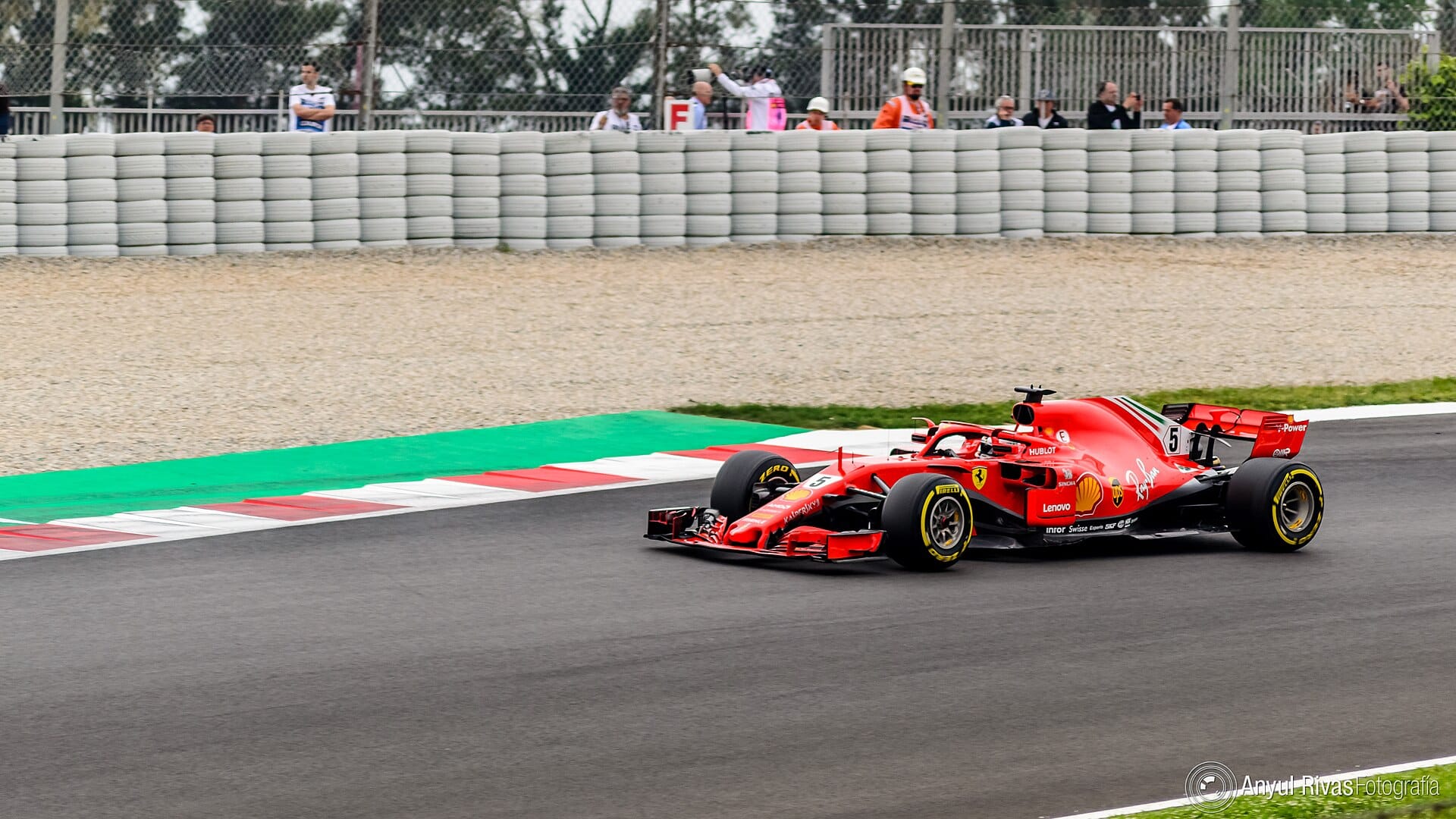 A red Ferrari Formula 1 racing car driving on a racetrack during a Grand Prix event, captured from a front three-quarters angle with spectators and barriers in the background.