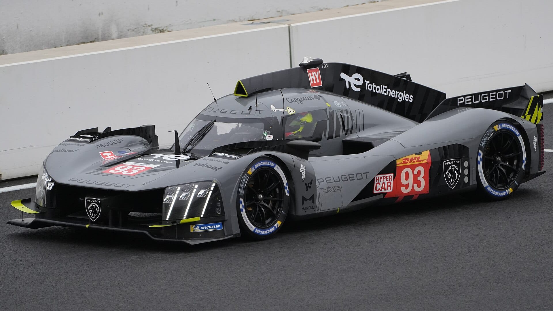 A grey Peugeot 9X8 race car with number 93 driving on a track alongside a barrier.