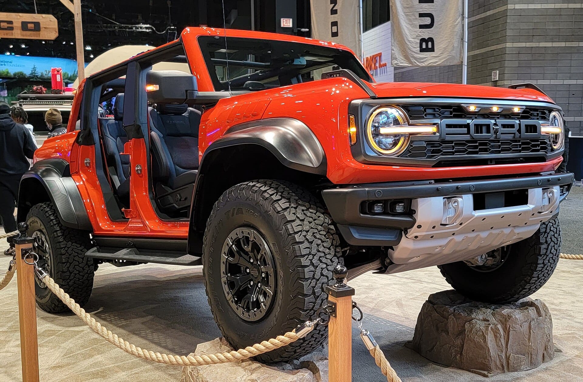 An orange Ford Bronco Raptor displayed indoors on an auto show floor, parked over artificial rocks with ropes cordoning off the exhibit.