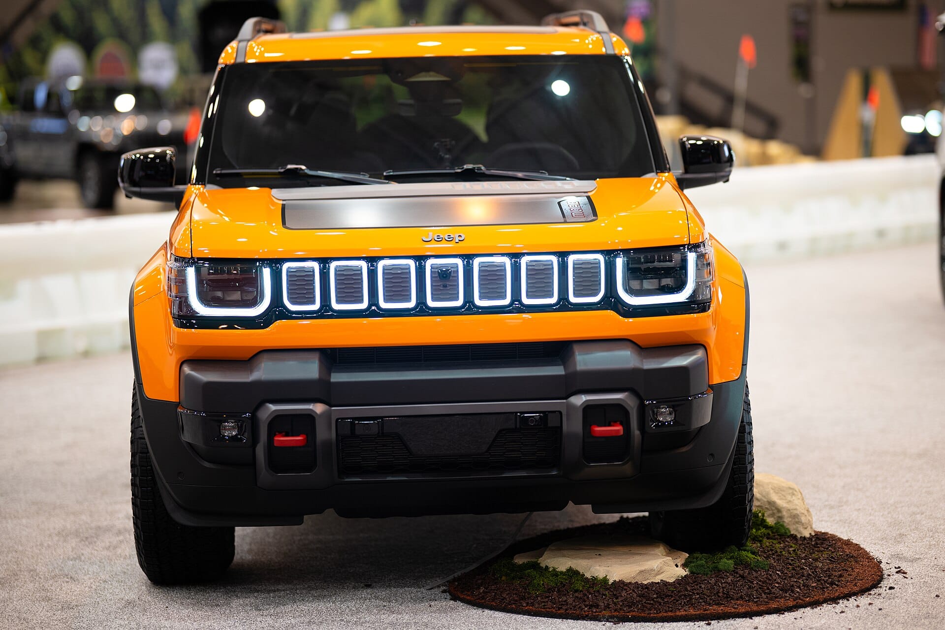 Frontvy av en orange Jeep Recon på en bilsalong inomhus, med upplyst grill och robust stötfångare.