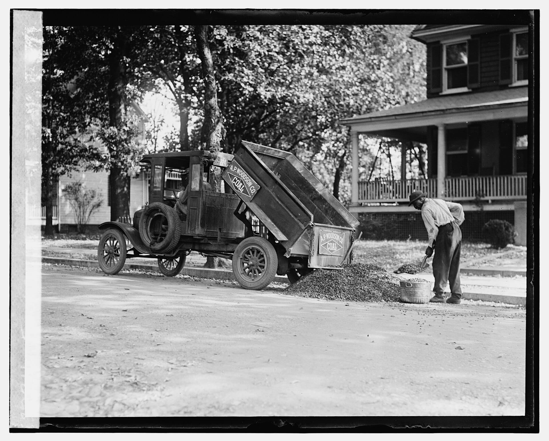 An old black and white photo showing a Ford truck dumping coal in front of a residential house with a worker shoveling.