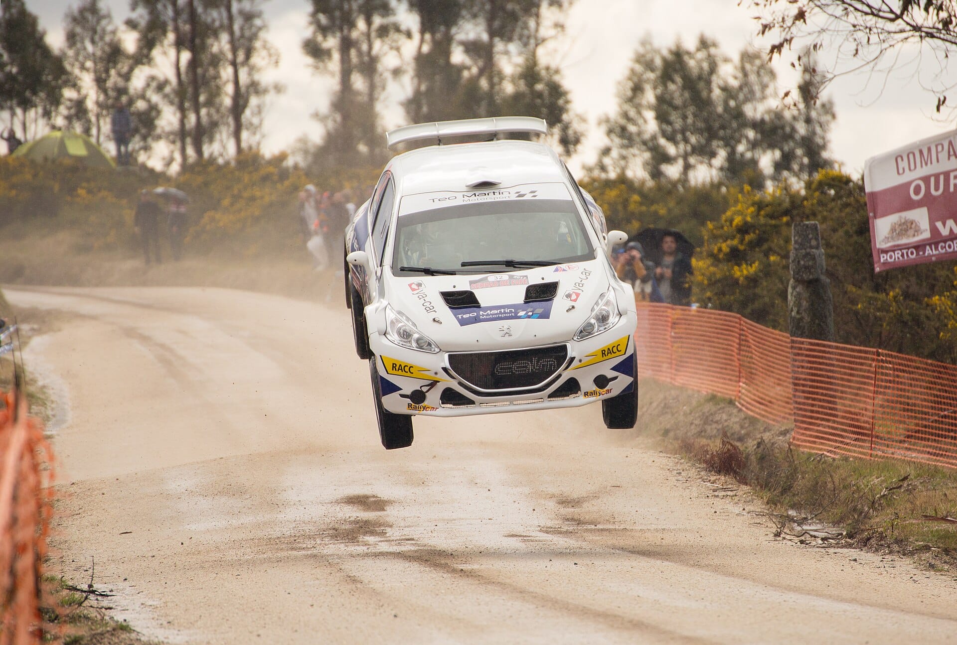 A rally car airborne after jumping over a crest on a dirt road during a rally event, with spectators watching from the sidelines.