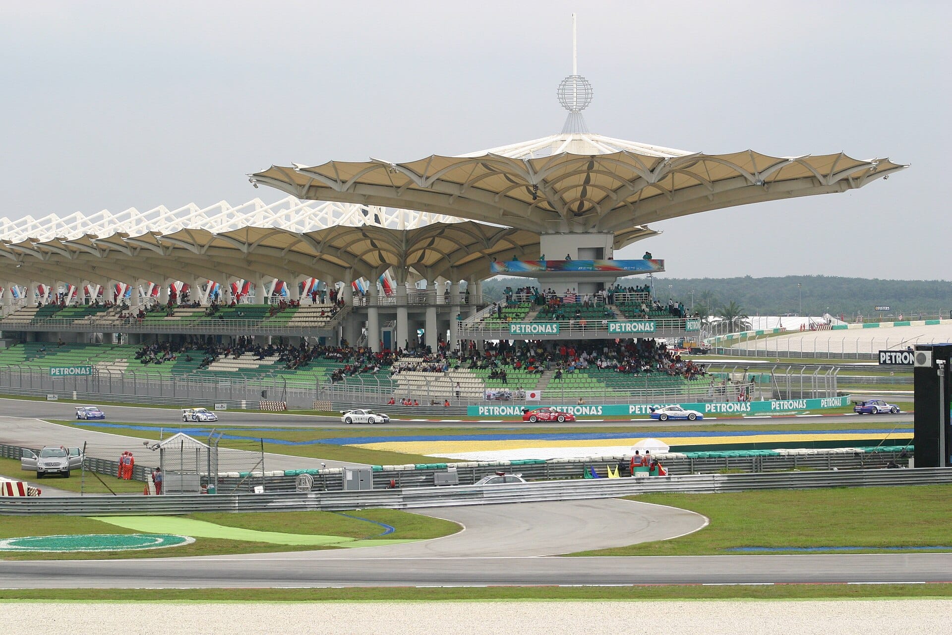 A wide shot of the Sepang International Circuit showing the grandstand with its distinctive umbrella-like roof structure, crowds of spectators, and racing cars on the track.