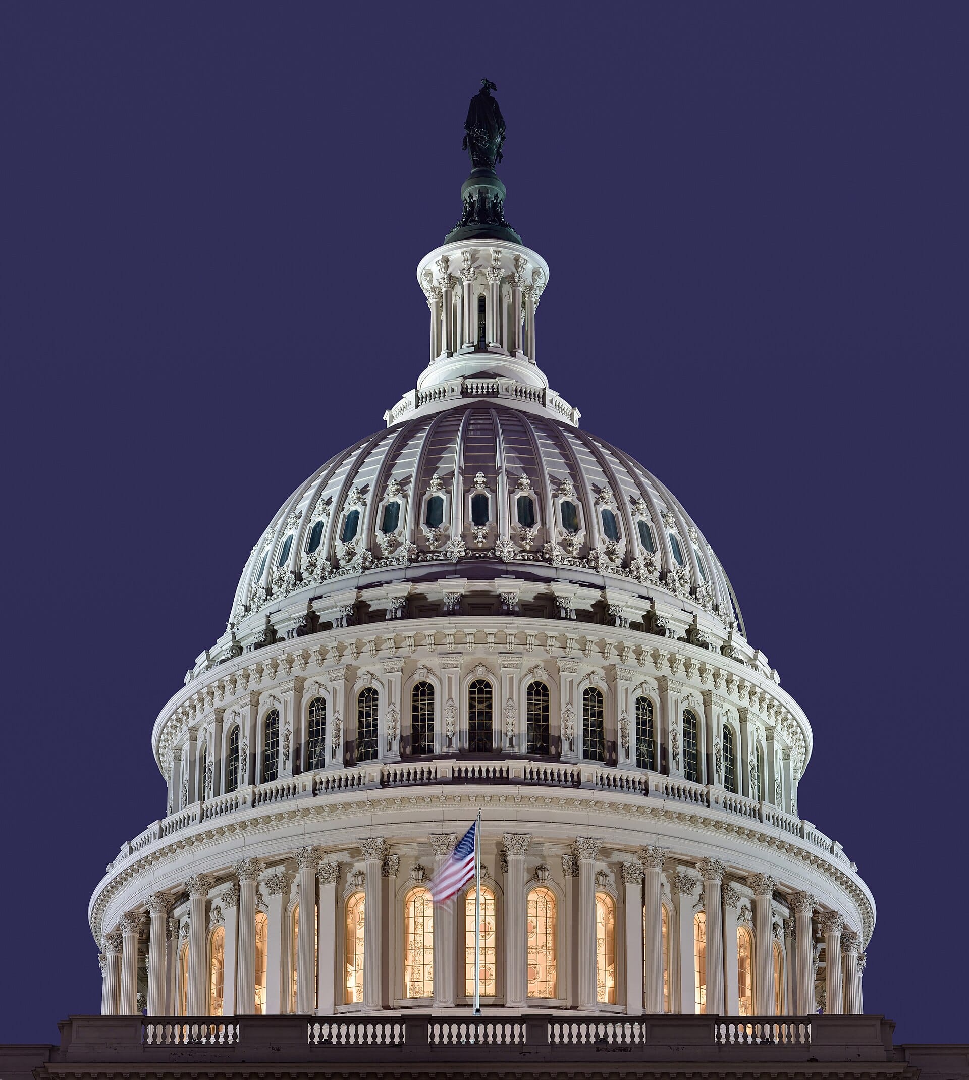 A low-angle, nighttime shot focusing on the illuminated dome of the United States Capitol in Washington, D.C., with an American flag flying on a pole in front of the colonnade against a deep purple evening sky.