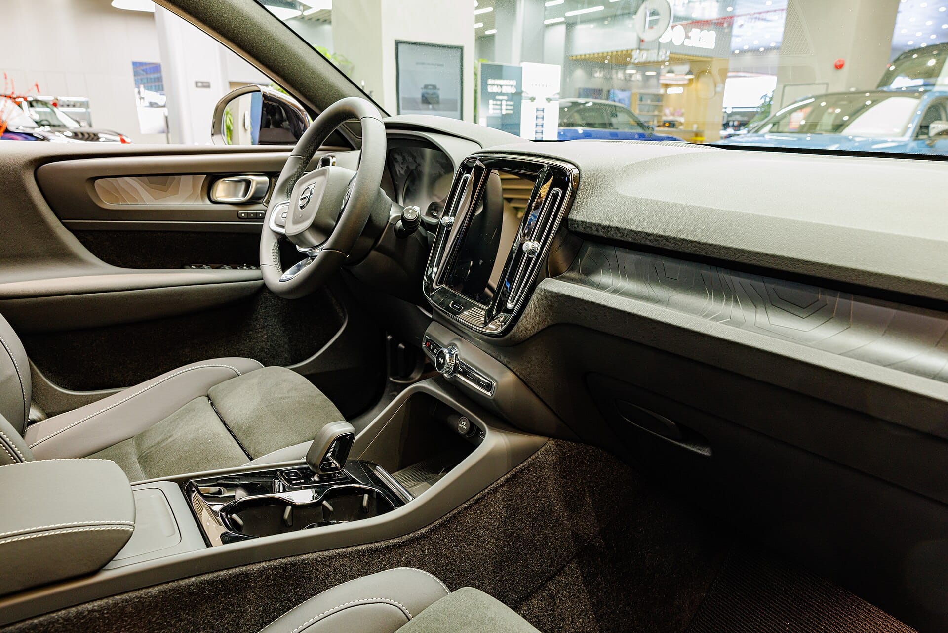 An eye-level interior view of a modern Volvo car dashboard, steering wheel, and front seats inside a showroom.