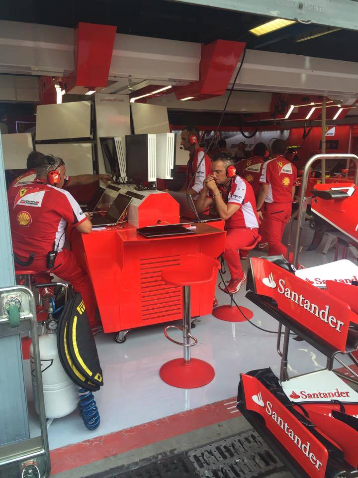 A view of the Ferrari pit lane garage during a race weekend, showing engineers and mechanics working at computer monitors surrounded by team equipment and red-and-white branding.