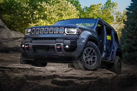 A dark blue Jeep Recon parked on rocky terrain with green trees in the background.