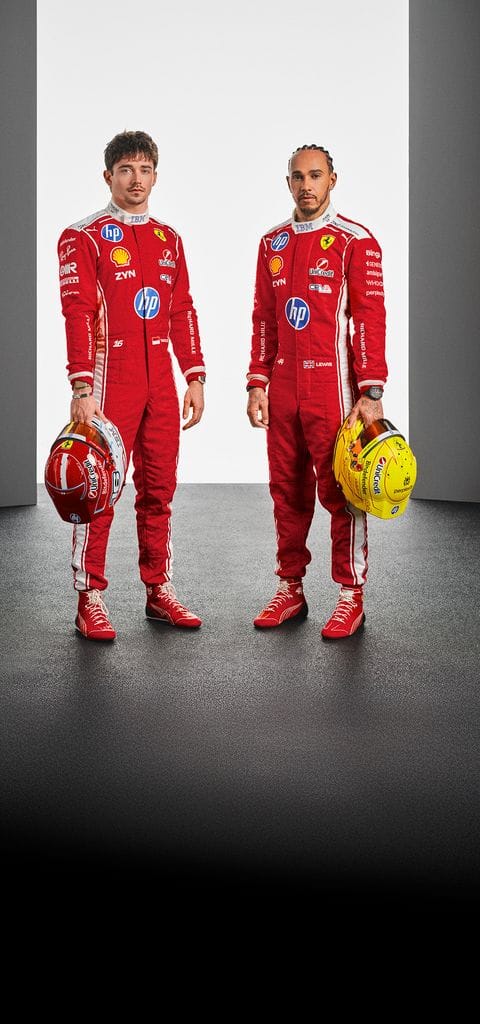 Full-length portrait of Charles Leclerc and Lewis Hamilton standing side-by-side in red Ferrari racing suits, holding their helmets in a studio setting.