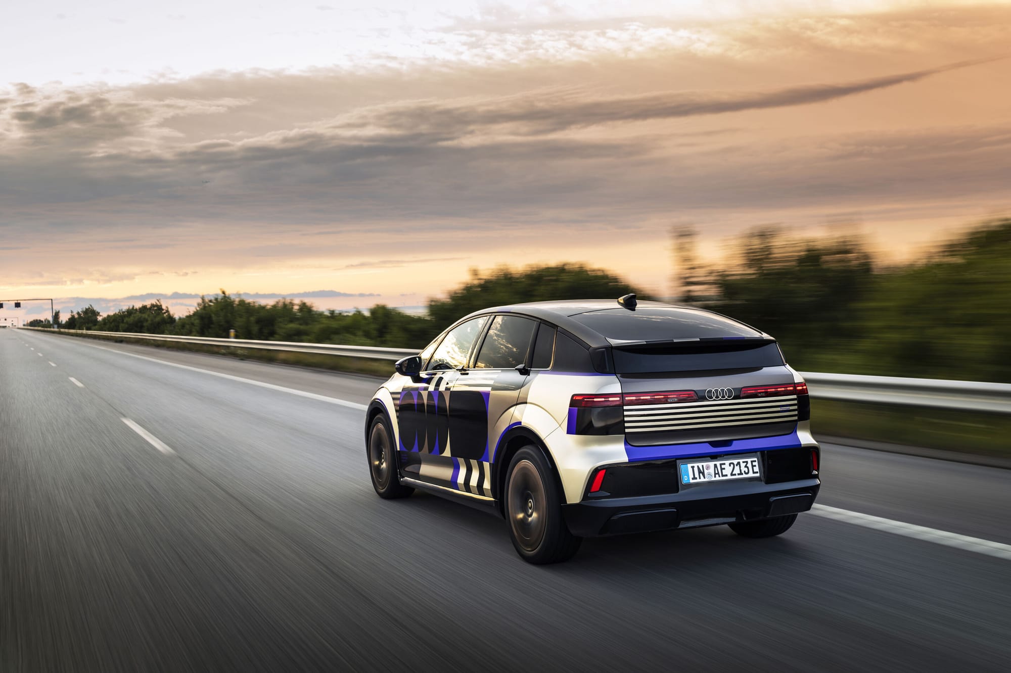 An Audi A2 E-Tron prototype driving on a highway during sunset, viewed from the rear three-quarters angle with motion blur on the road and surroundings.