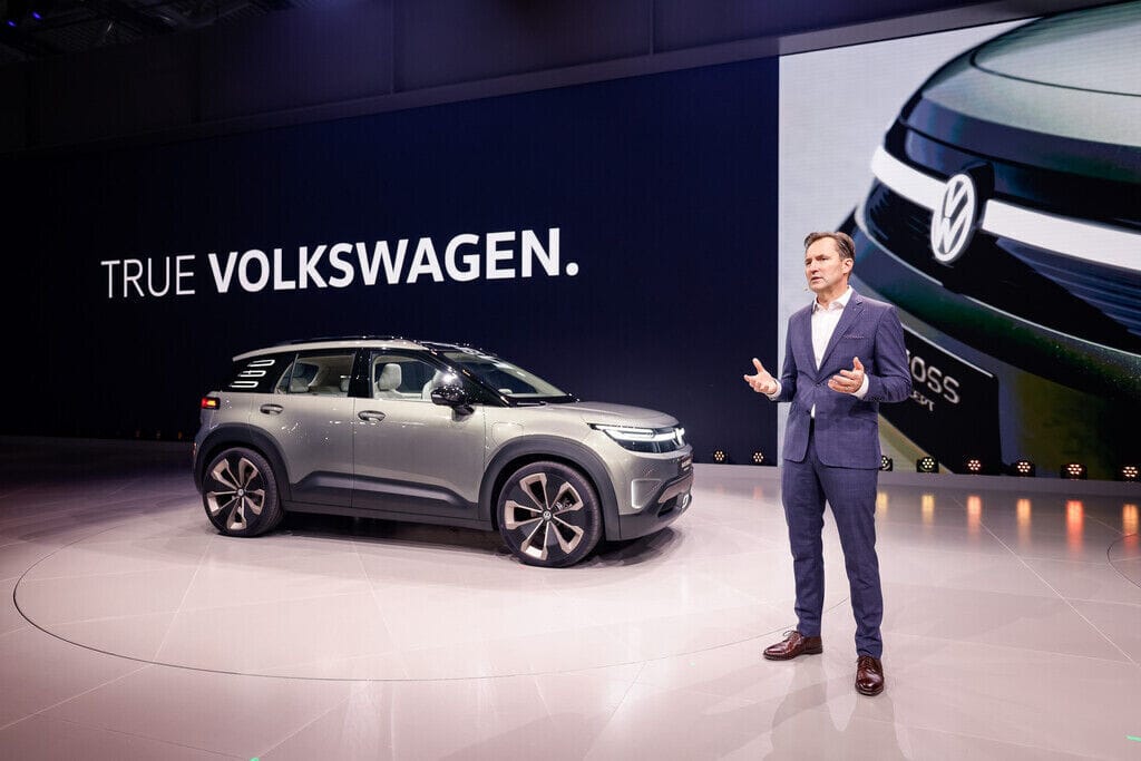 A presenter on a stage unveiling a new Volkswagen concept car with the text 'TRUE VOLKSWAGEN' on the backdrop.