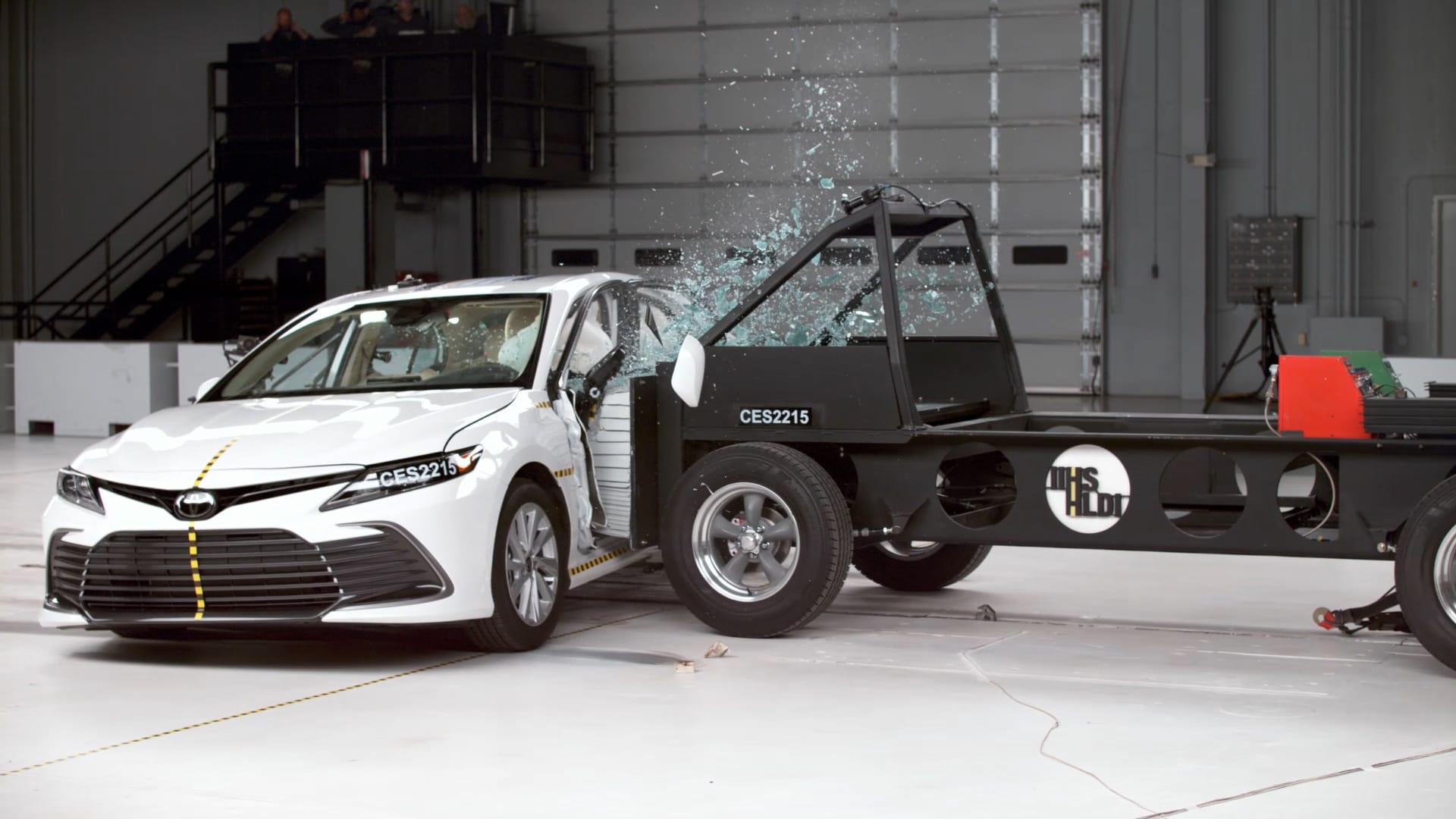 A white car undergoing a side crash test in a laboratory setting with IIHS branding visible on the barrier cart.