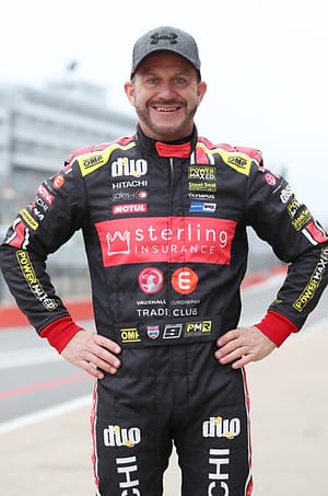 A portrait of race car driver Rob Collard wearing his racing suit and a baseball cap, standing outdoors near a racetrack.