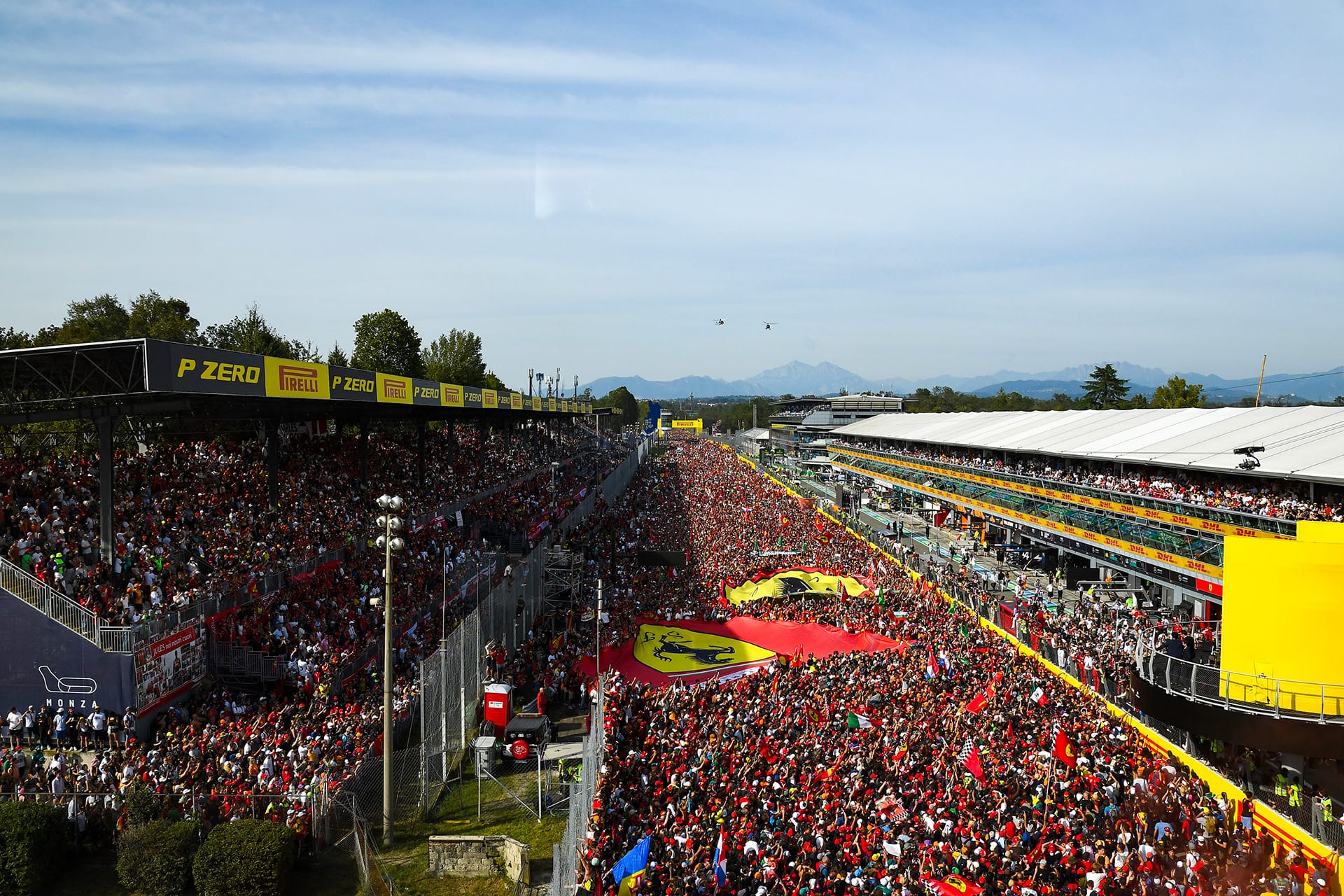A high-angle wide establishing shot of the Italian Grand Prix at Monza, showing a massive crowd of fans filling the racetrack and grandstands with flags and banners.