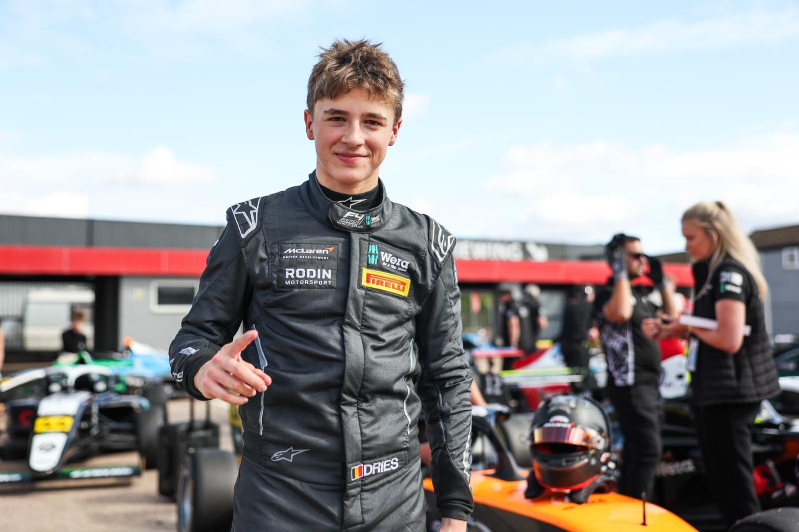 A young male racing driver standing in a paddock, wearing a dark racing suit with sponsor logos, pointing towards the camera with other people and race cars in the background.