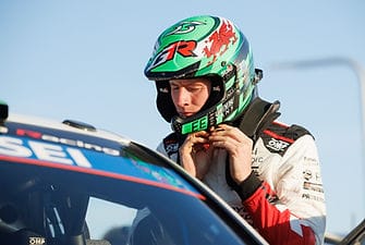 A portrait of rally driver Elfyn Evans wearing a racing suit and a green, black, and red helmet while preparing to enter his rally car.