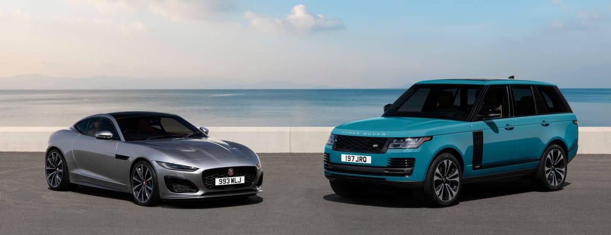 Two Jaguar Land Rover vehicles, a silver Jaguar sports car and a teal Range Rover SUV, parked side by side by the coast.
