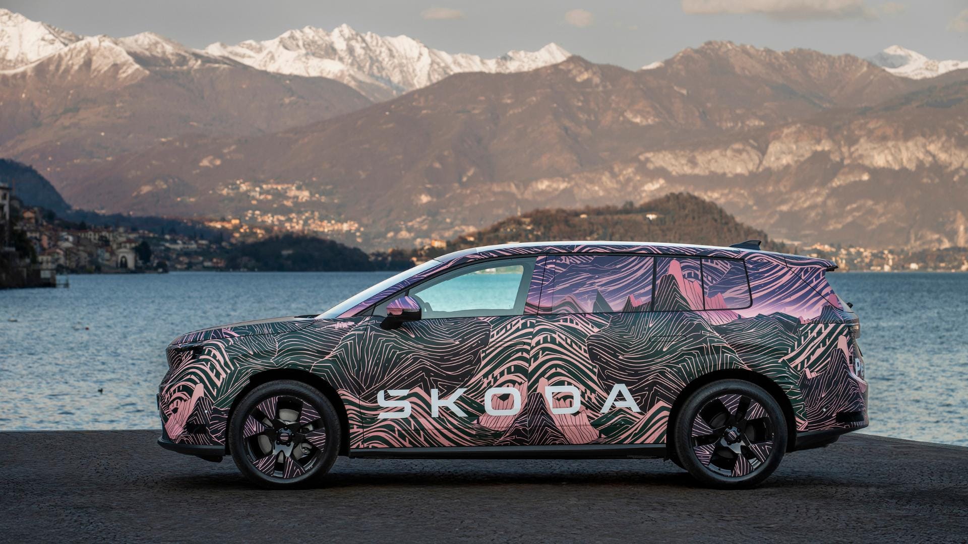 A side profile view of a Skoda SUV wrapped in camouflage featuring pink and green mountain patterns, parked near a lake with snowy mountains in the background.