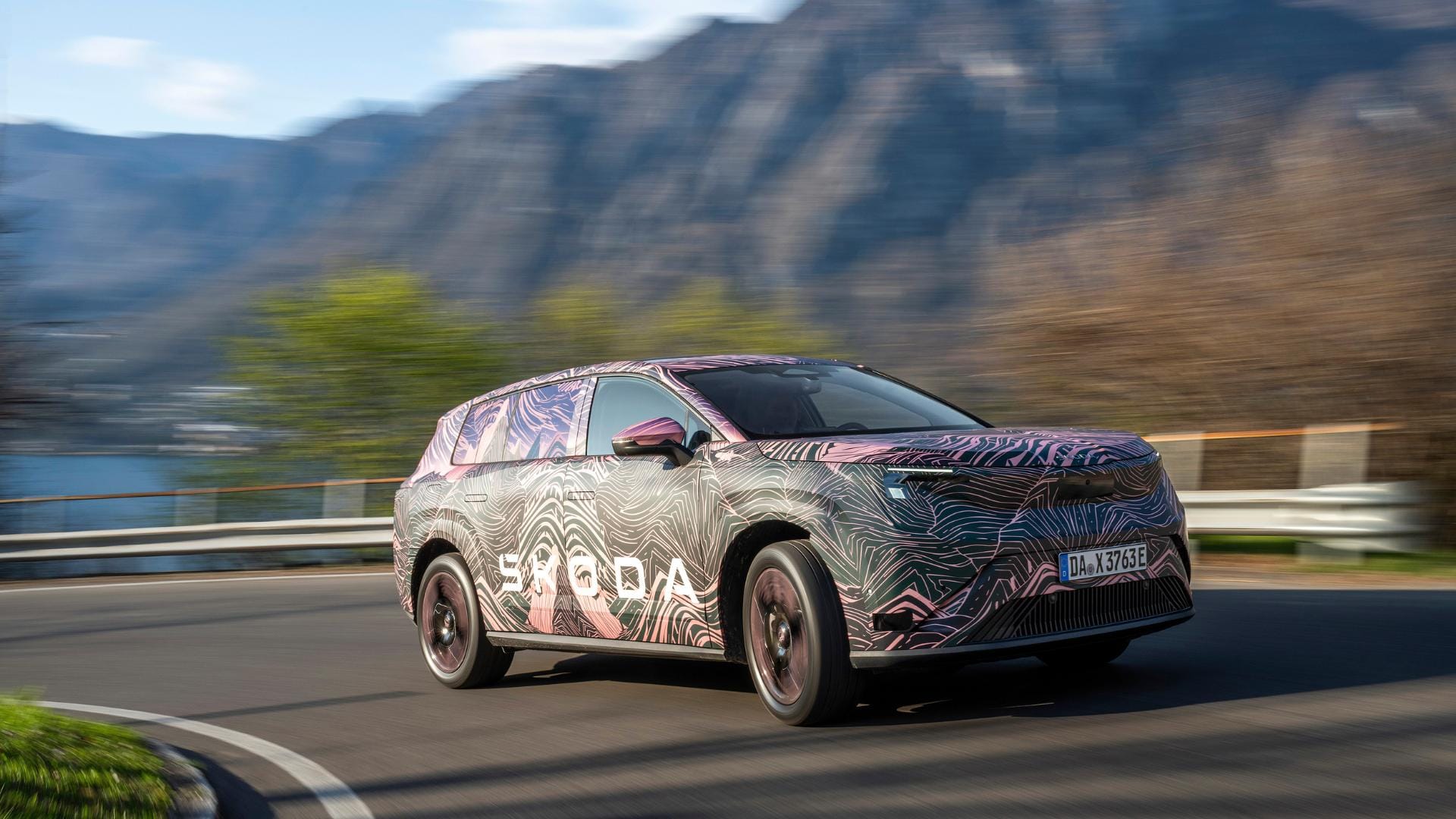 A camouflaged Skoda prototype vehicle driving on a winding coastal road with mountains in the background, captured in motion with motion blur in the background.
