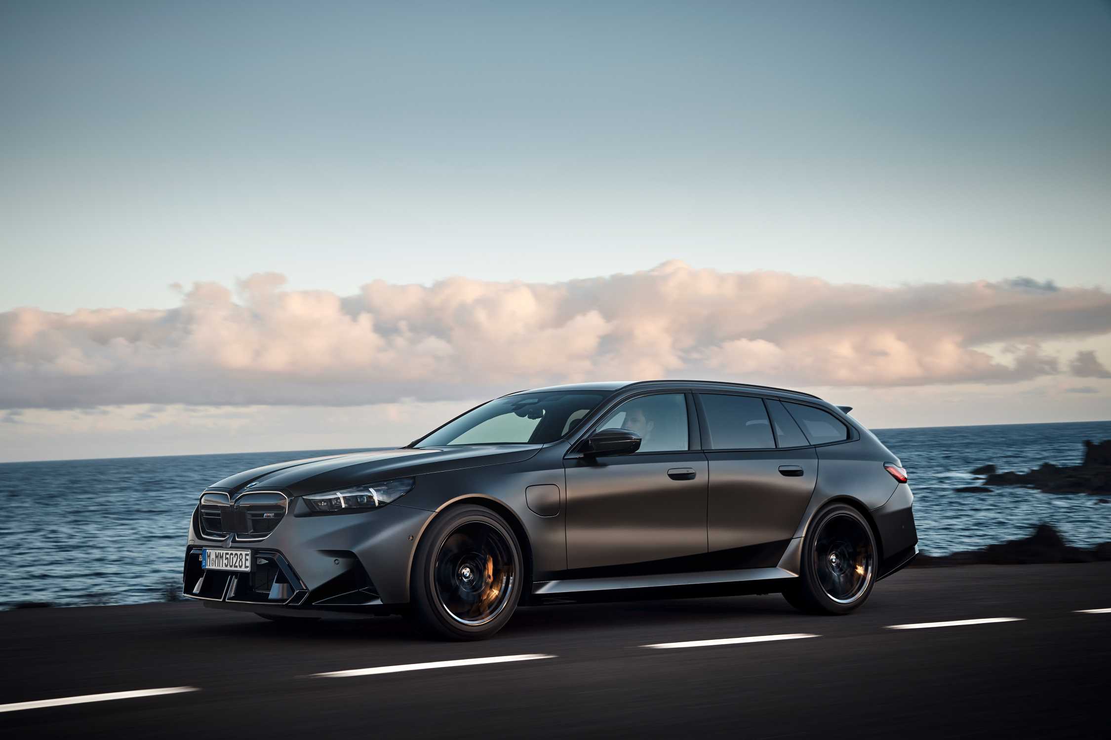 A dark grey BMW M5 Touring is driving on a coastal road with the sea and cloudy sky in the background, captured in a dynamic promotional shot.