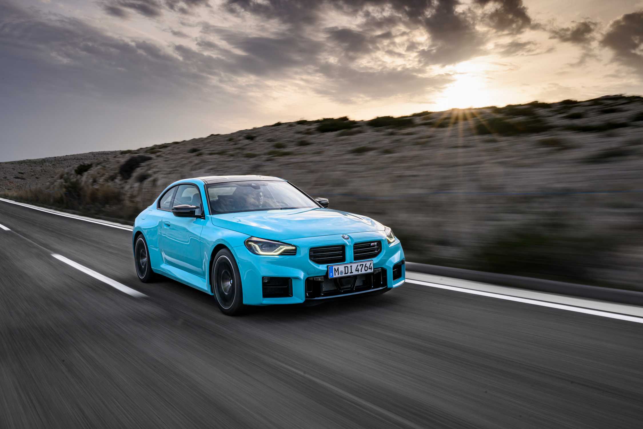 A light blue BMW M2 driving on a coastal road during sunset, captured in motion with motion blur on the road.