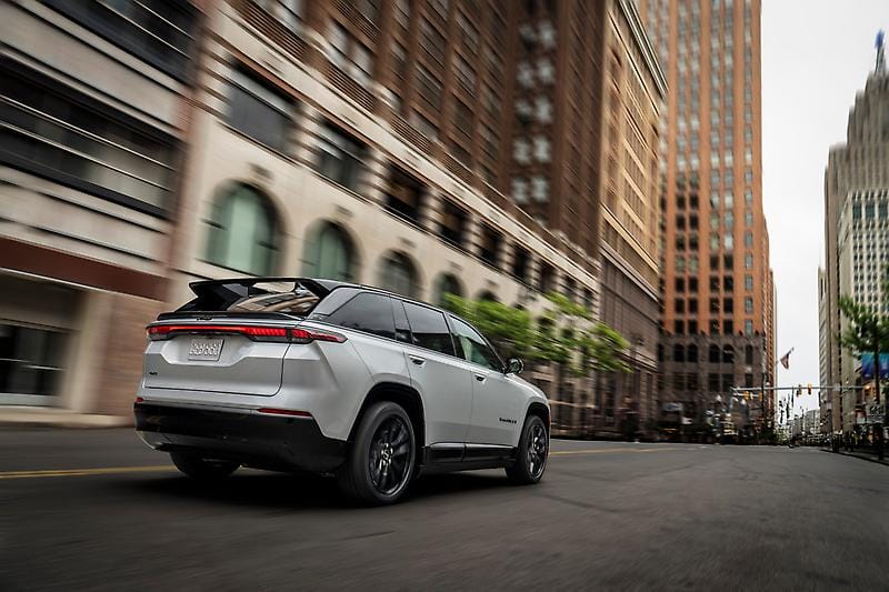 A white Jeep Grand Cherokee driving down a city street flanked by tall buildings with motion blur suggesting speed.