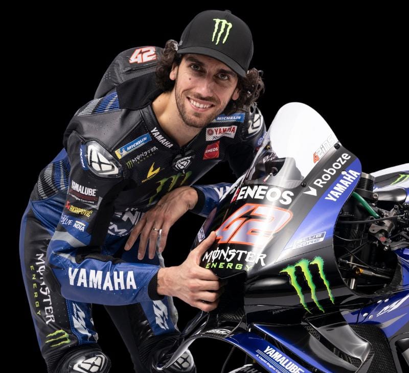 Motorcycle racer Alex Rins smiling while posing next to his Yamaha race motorcycle in a studio setting.