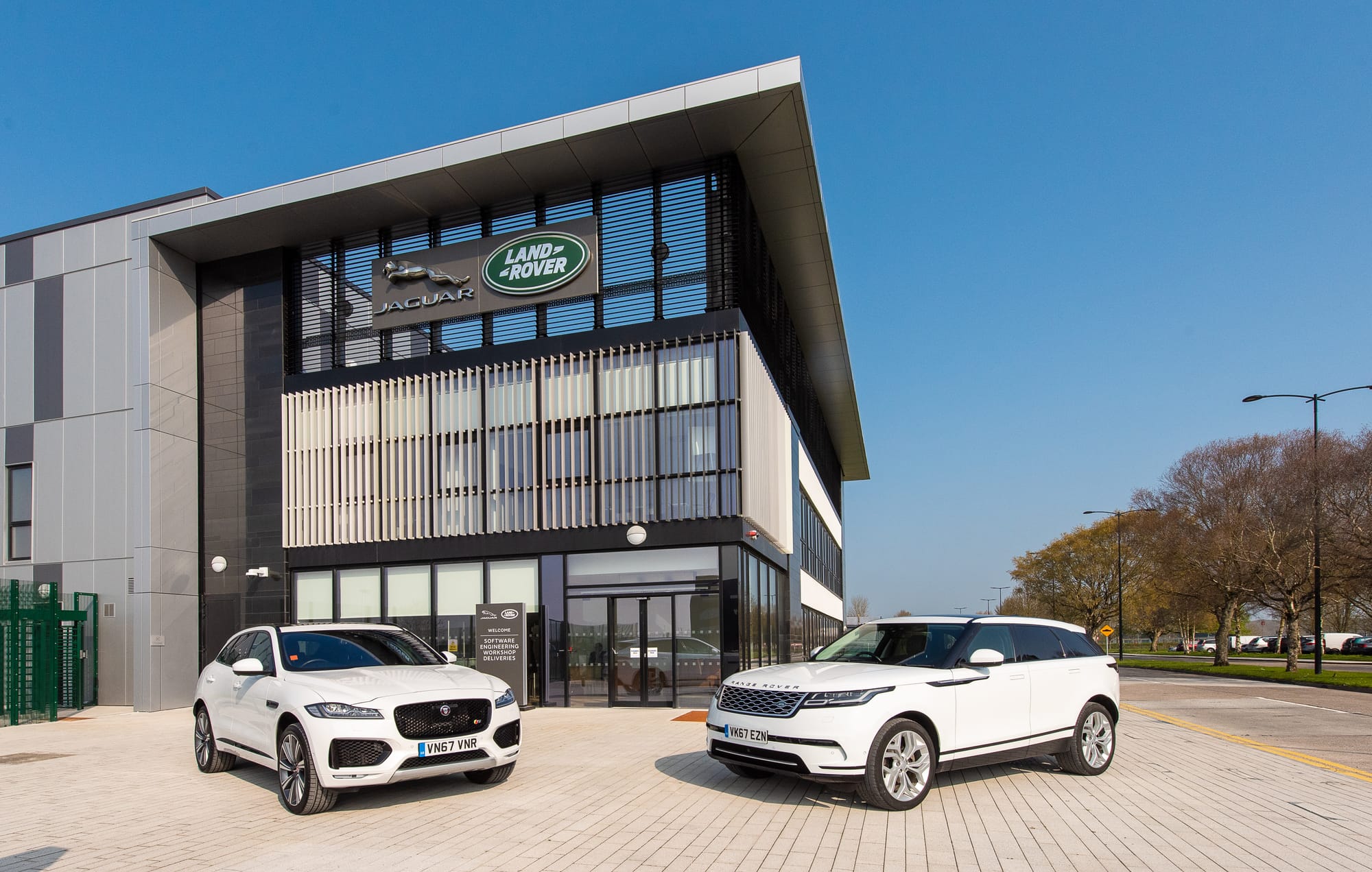 An exterior view of a modern Jaguar Land Rover dealership building with two white vehicles parked in the front plaza under a clear blue sky.