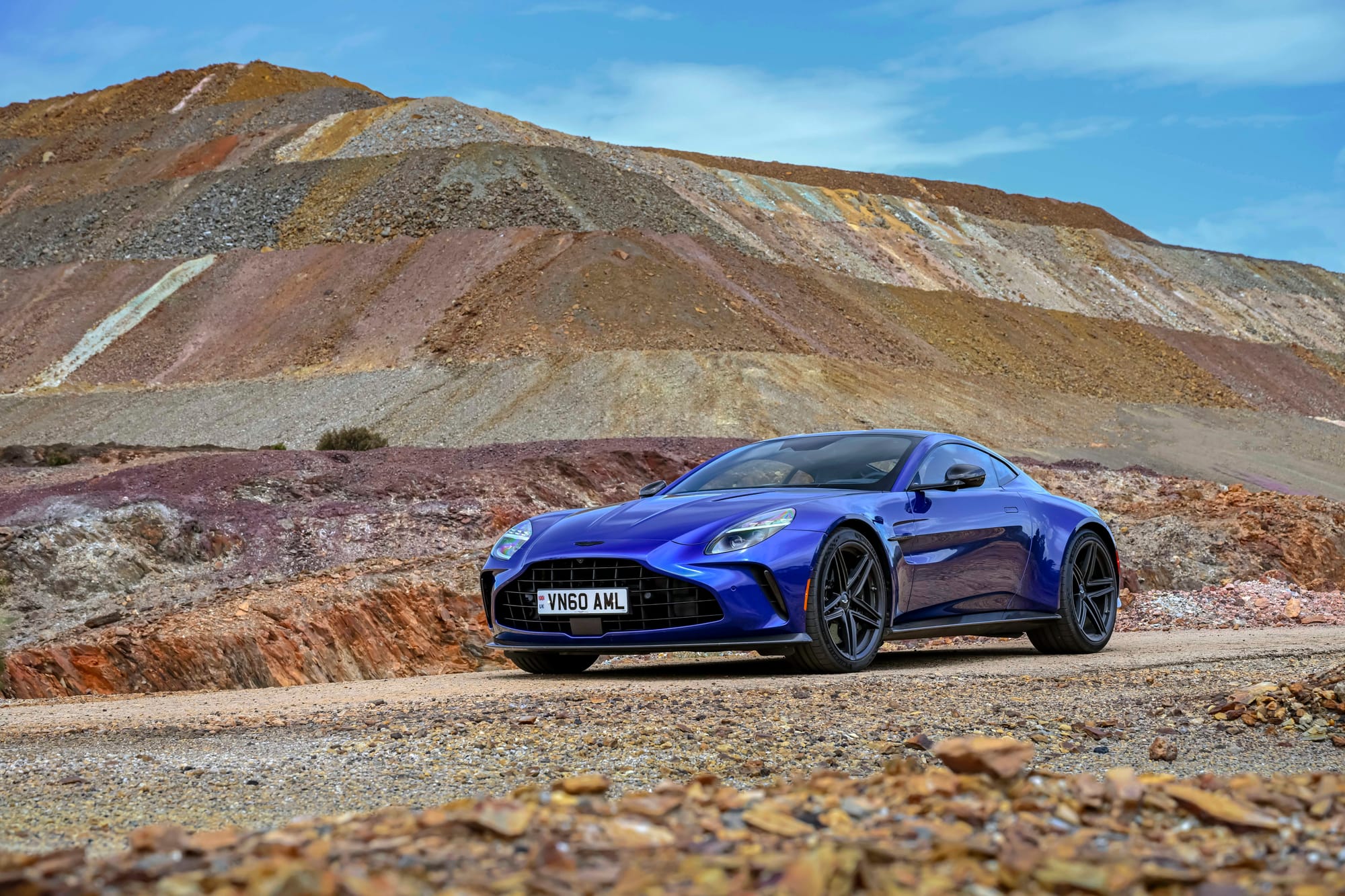 A dark blue Aston Martin Vantage sports car parked on a gravel path against a backdrop of colorful, layered rocky hills.