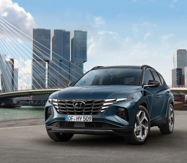 A modern blue Hyundai Tucson parked outdoors near a waterfront with a bridge and skyscrapers in the background.