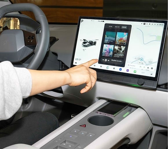 A person interacting with the Pleos Infotainment System touchscreen inside a modern car interior, showing maps and media controls.