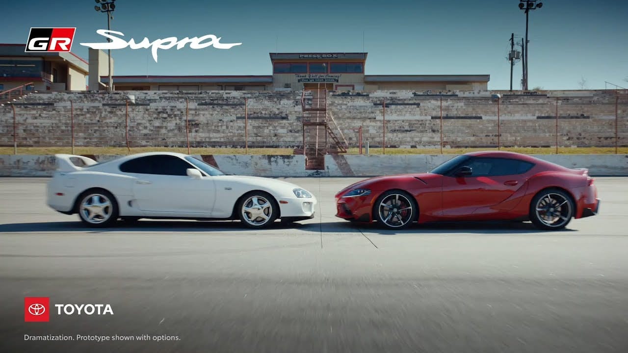 Two generations of Toyota Supras, an older white model and a newer red GR Supra, parked facing each other on a racetrack with a GR Supra logo overlay in the top left corner.