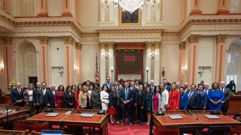 A large group of legislators standing together inside the California State Senate chamber.
