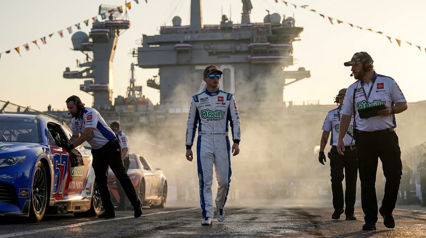 En racerförare i vit overall och solglasögon går framåt på pitlane framför ett hangarfartyg, flankerad av funktionärer och en blå racerbil.