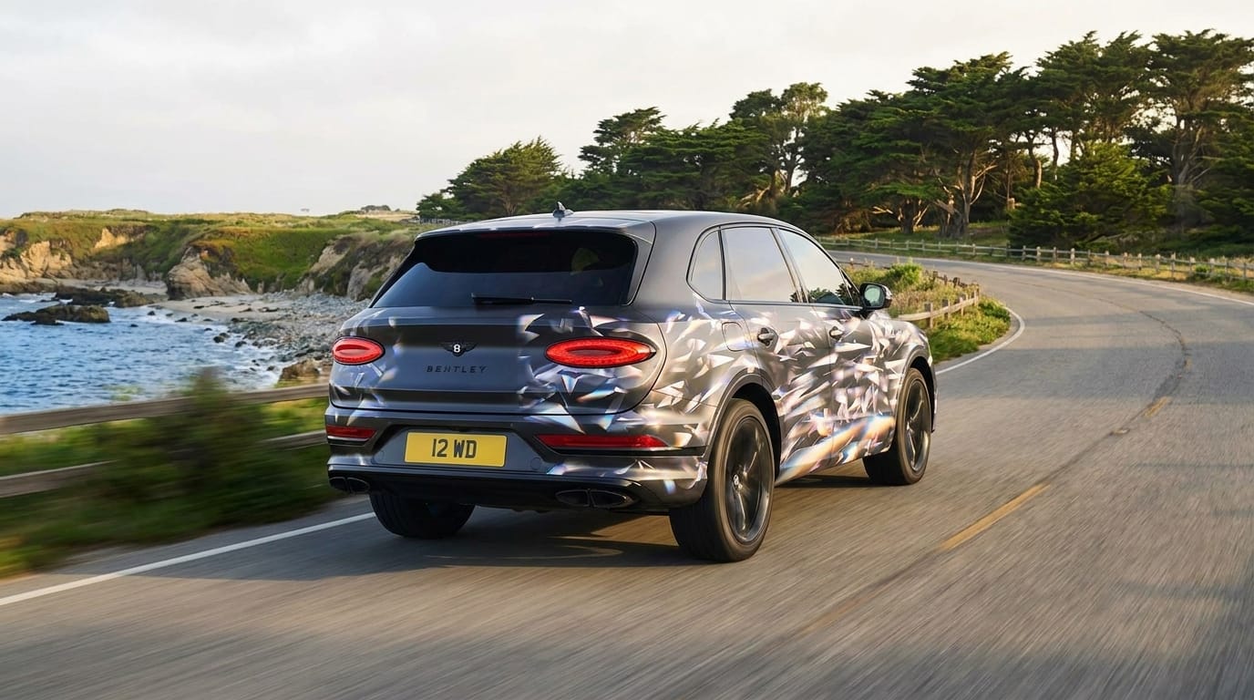 Mörk SUV med geometrisk kamouflage kör på en kurvig kustväg bredvid havet och gröna träd.