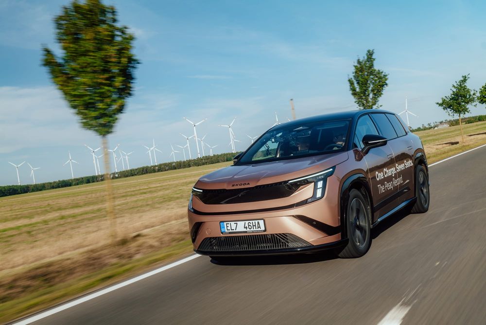 A bronze Skoda SUV driving on an asphalt road in a rural landscape with wind turbines in the background.
