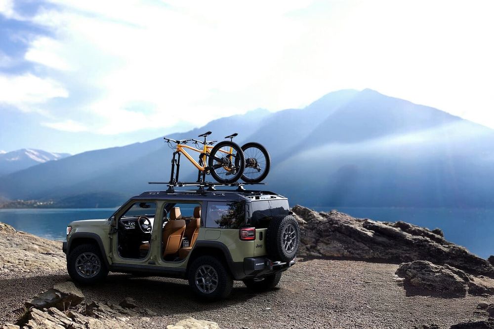 A green Jeep Recon parked on a rocky cliff overlooking a lake and mountains, with two bicycles mounted on the roof rack.