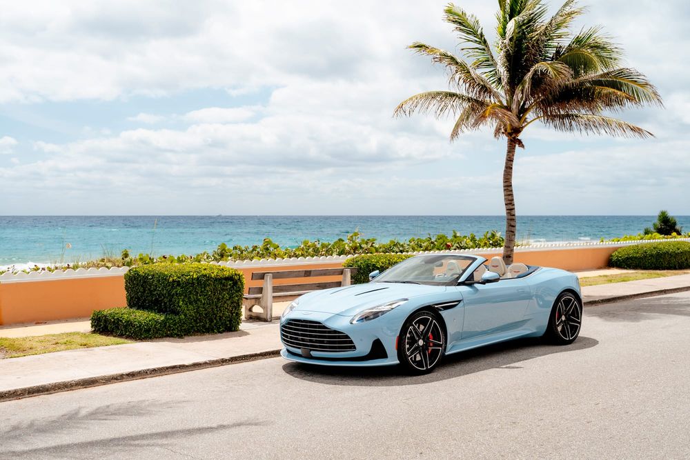 A light blue Aston Martin convertible parked along a scenic coastal road with a palm tree and the ocean in the background.