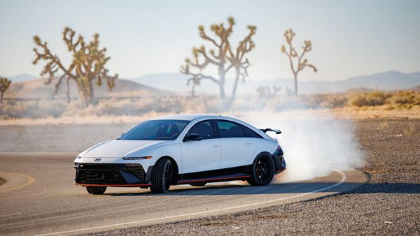 A white Hyundai performance electric sedan drifting on an asphalt track in a desert landscape with smoke billowing from its tires and Joshua trees in the background.
