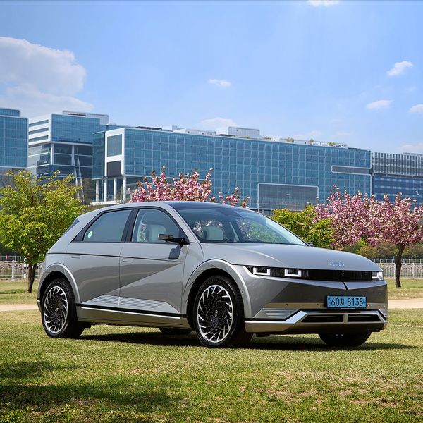 A silver Hyundai Ioniq 5 electric vehicle parked on a green lawn in front of modern glass office buildings and blooming trees.