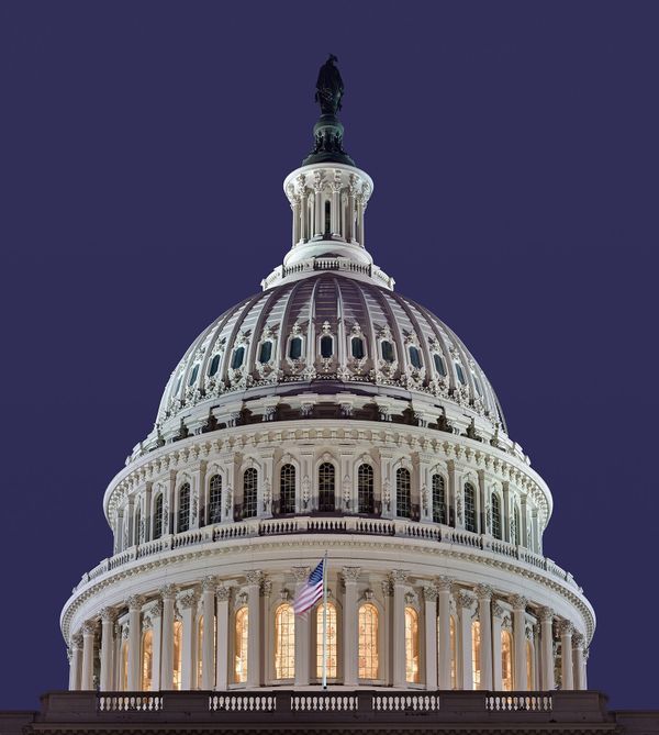 A low-angle, nighttime shot focusing on the illuminated dome of the United States Capitol in Washington, D.C., with an American flag flying on a pole in front of the colonnade against a deep 