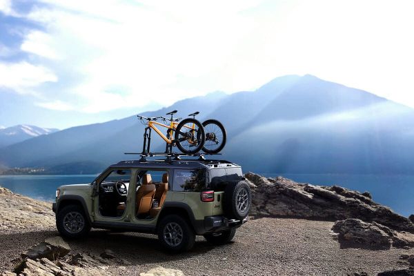 A green Jeep Recon parked on a rocky cliff overlooking a lake and mountains, with two bicycles mounted on the roof rack.