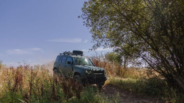 An off-road dark green vehicle driving on a dusty dirt path surrounded by tall grass and bushes under a clear sky.