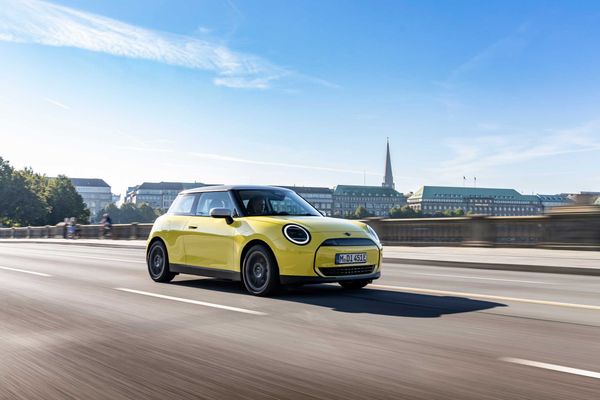 A yellow Mini Cooper Electric driving along an open city bridge with classic buildings and a clear blue sky in the background.