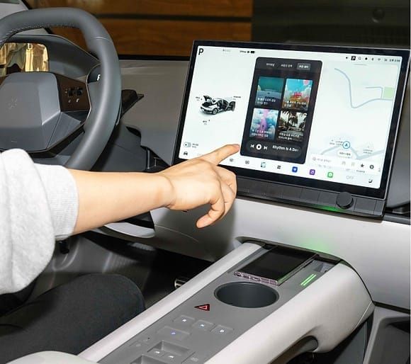 A person interacting with the Pleos Infotainment System touchscreen inside a modern car interior, showing maps and media controls.