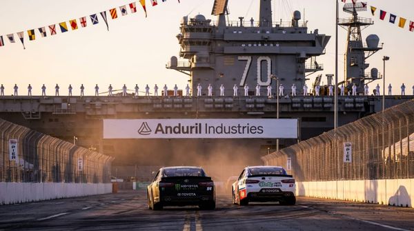 Bakifrånfoto av två stock race-bilar som kör sida vid sida på en bana bredvid ett hangarfartyg i solnedgång.